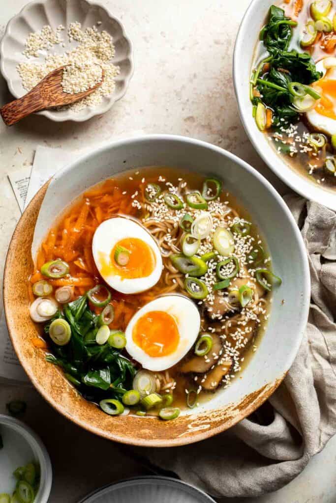 Delicious homemade vegan ramen with carrots, spring onions, noodles, egg and mushroom served in a white bowl topped with sesame seeds.