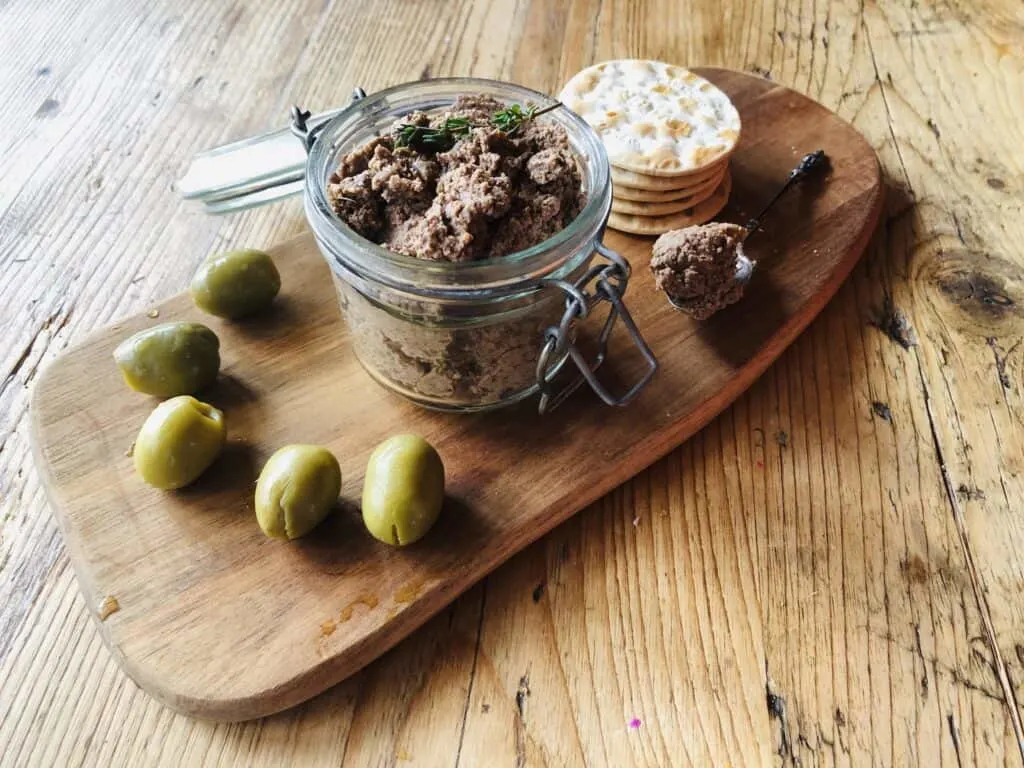 homemade vegan mushroom pate in a jar, on a chopping board, with crackers and fresh olives.