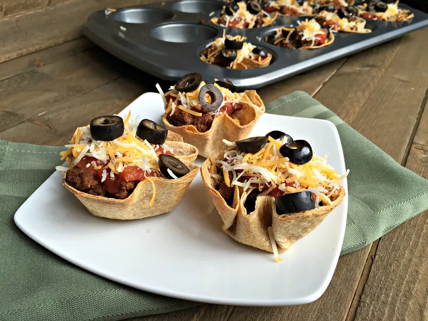 upclose beef taco cups topped with olives and grated cheese on a white plate on a table with a baking tray in the background with beef taco cups