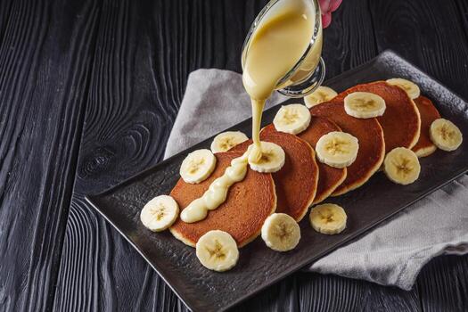 Delicious homemade vegan banana pancakes topped with thin sliced fresh banana with pouring custard served on a black plate