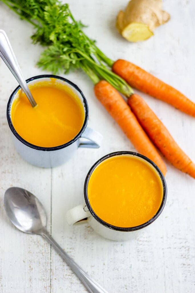2 cups of creamy homemade carrot and ginger soup on table with fresh carrots and fresh ginger.