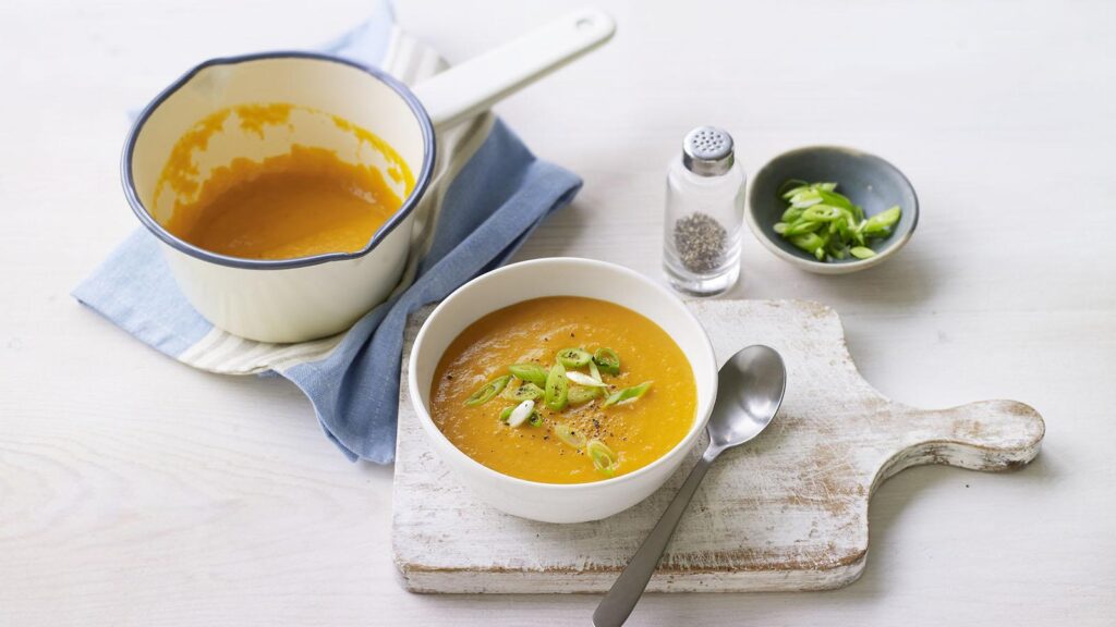 creamy homemade carrot and ginger soup with black pepper served in a white bowl and placed on top of a chopping board with a tablespoon on the side.